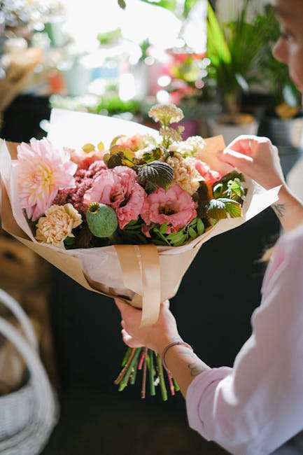 A person holding a beautifully arranged floral bouquet featuring pink peonies, cream carnations, and small white filler flowers, complemented by dark green leaves. The bouquet is wrapped in beige and 