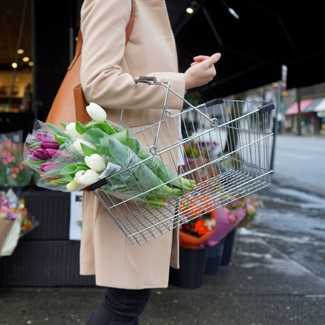 A person wearing a beige coat holding a wire basket containing a bouquet of fresh tulips, predominantly white and purple, wrapped in clear plastic with green foliage visible. The basket is suspended o