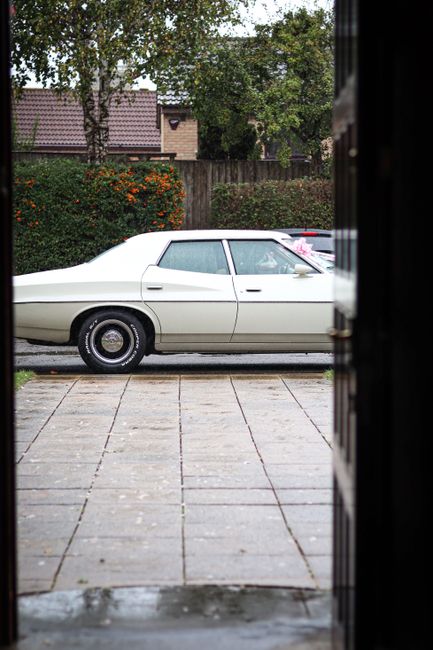 A classic white vintage car parked outside on a paved driveway, viewed from an open doorway or window. The car features sleek, smooth lines with black tires and shiny hubcaps. In the background, there