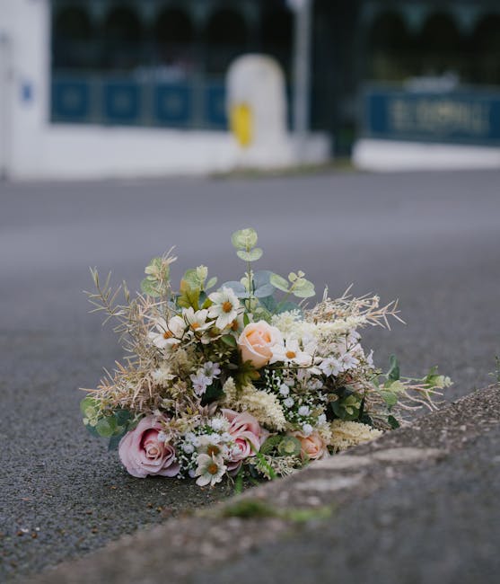 A delicate floral bouquet resting on an asphalt pavement beside a curb, featuring soft pink roses, white daisies, and cream-colored blossoms complemented by eucalyptus leaves and wispy, dried foliage.
