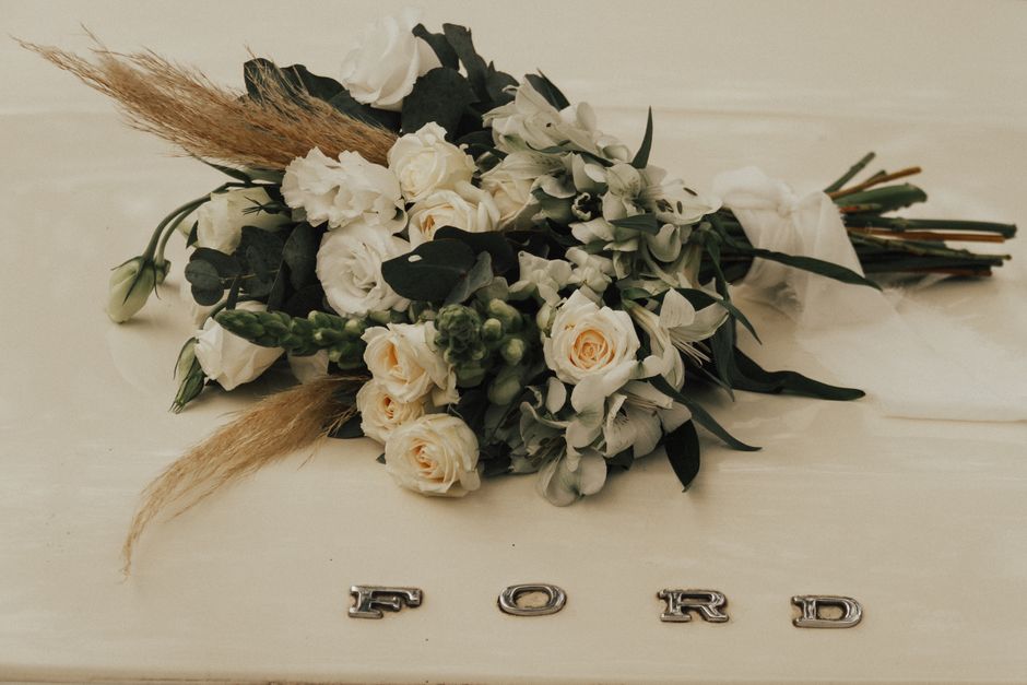 A floral bouquet arrangement featuring cream and white roses, white lisianthus, and green foliage, including eucalyptus leaves and buds, with accents of dried pampas grass. The flowers are arranged in