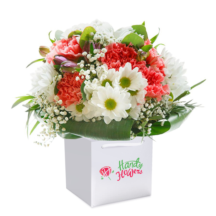 Poised within a clean white square gift bag and photographed against a luminous white backdrop, this vibrant bouquet captures the spirit of Old Ford floristry. The composition springs to life with plush coral-pink and ruby-red carnations, their finely ruffled petals offering a dynamic, tactile richness at the heart of the display. White daisy-like chrysanthemums punctuate the arrangement, their bold lime-green centers drawing the eye and invoking the effortless freshness of Bow Common's green spaces. Graceful sprigs of dainty white baby's breath float among the larger flowers, infusing the overall arrangement with soft, cloud-like accents that balance its energetic core. On the left, several closed buds brushed with purple and blush pink add textural intrigue, hinting at hidden stories and future blooms. Framing the entire bouquet, lush, broad leaves in deep emerald tones form a glossy collar, providing both structure and a connection to the verdant parks and gardens near Old Ford Road. The sturdy ribbon handles of the minimalist white bag increase both function and elegance, ensuring the bouquet arrives in perfect condition anywhere in E3. Flooded with soft, even lighting, every floral detail is carefully rendered-an ideal visual for an article on local flower delivery, thank-you gifts, or thoughtful gestures that mark life's special occasions in Old Ford and surrounding communities.